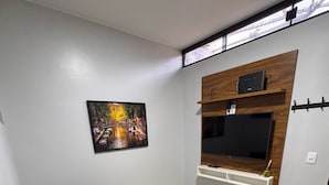 A living room with a black sofa, a mounted TV, a picture of a bridge, and a window with blinds.