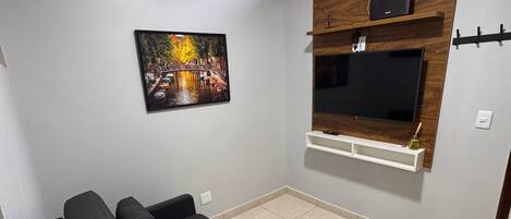A living room with a black sofa, a mounted TV, a picture of a bridge, and a window with blinds.