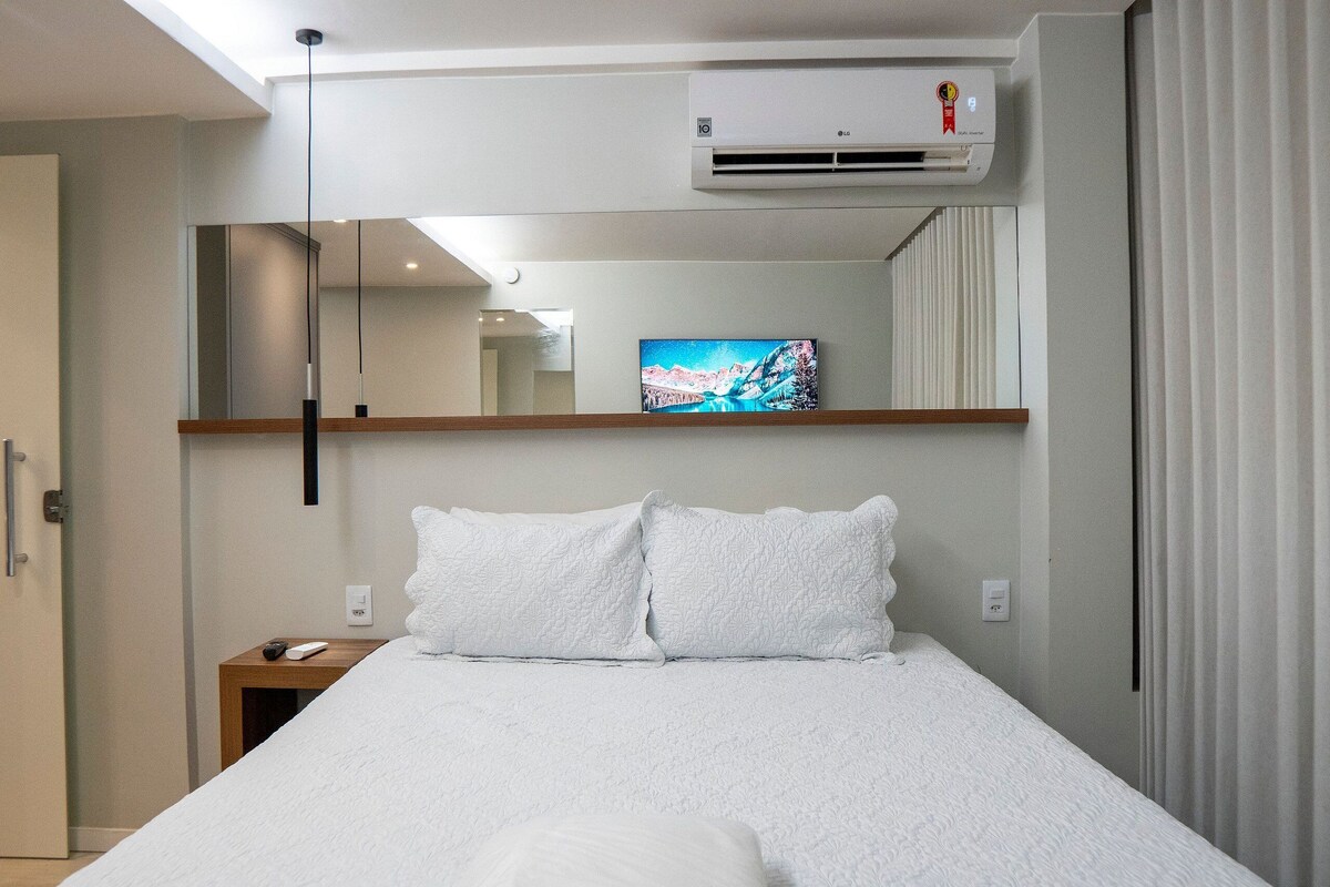 A hotel room with a bed, a mounted air conditioner, a painting on the wall, and a bedside table with a remote control.