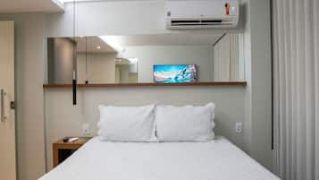 Una habitación de hotel con cama, aire acondicionado instalado, un cuadro en la pared y una mesita de noche con control remoto.