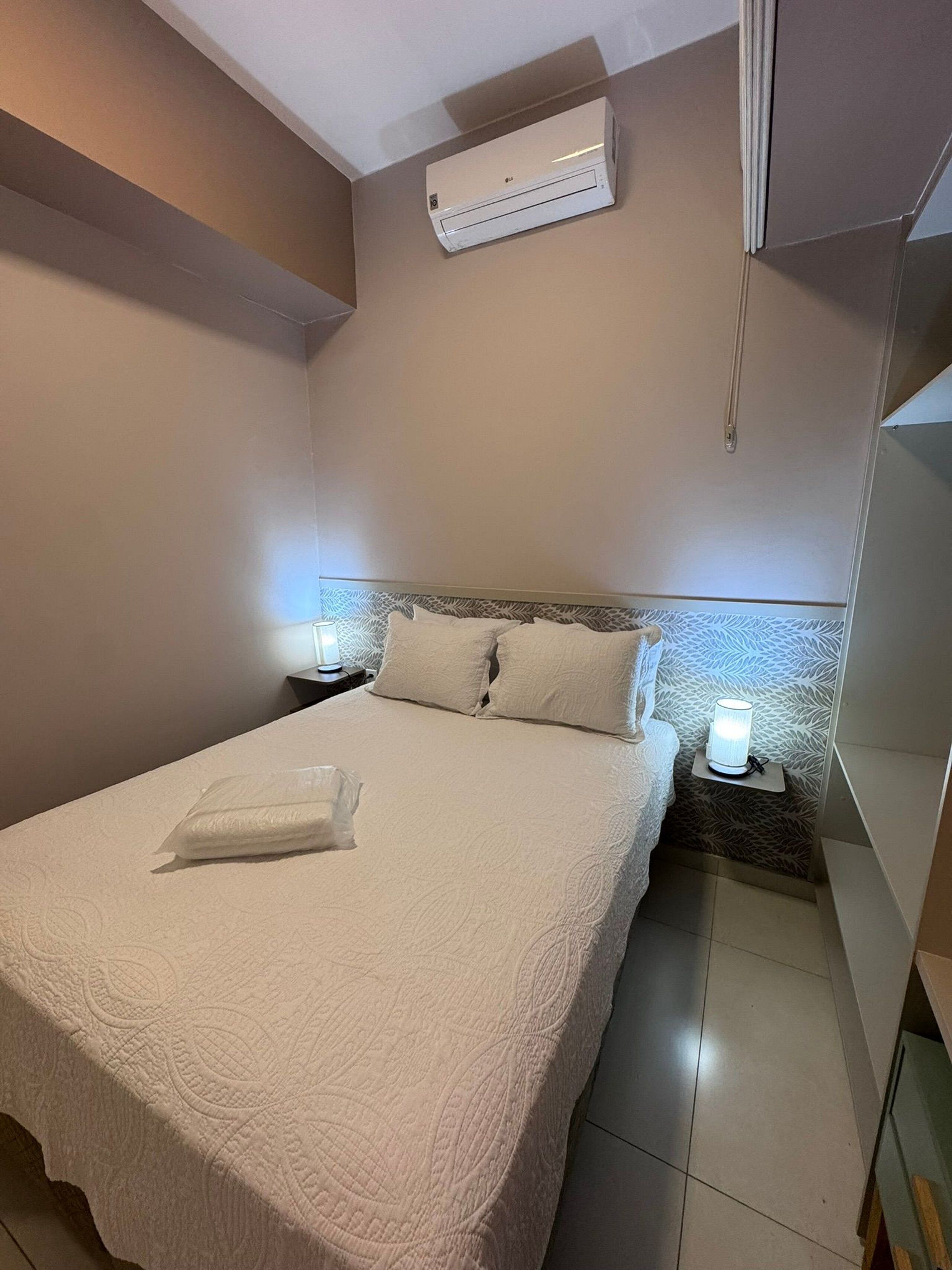 A neatly made bed with a white comforter and pillows, a bedside lamp, and an air conditioning unit mounted on the wall.