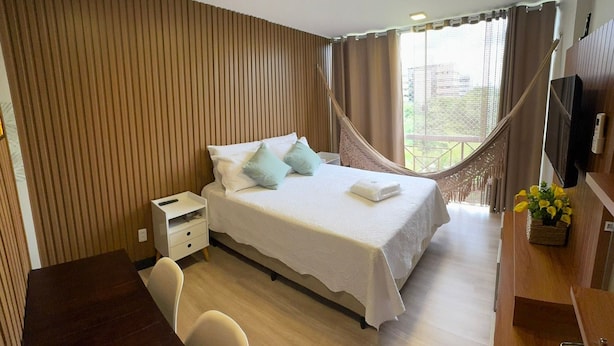 A hotel room with a bed, bedside table, chair, desk, TV, and a hammock.