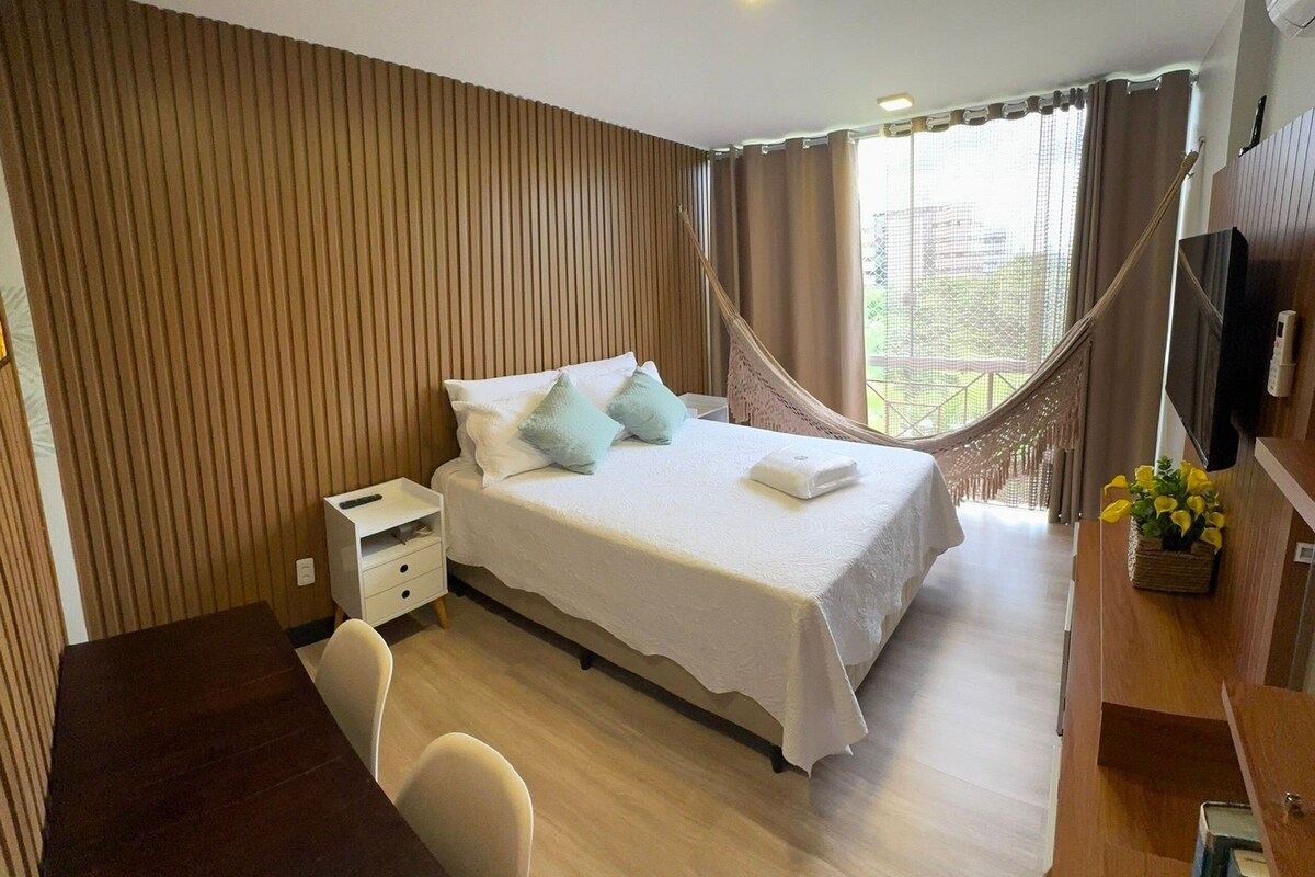 A hotel room with a bed, bedside table, chair, desk, TV, and a hammock.