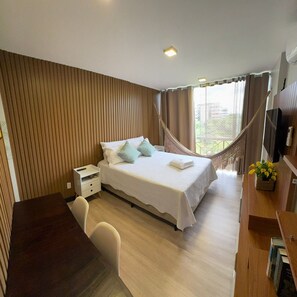 A hotel room with a bed, bedside table, chair, desk, TV, and a hammock.
