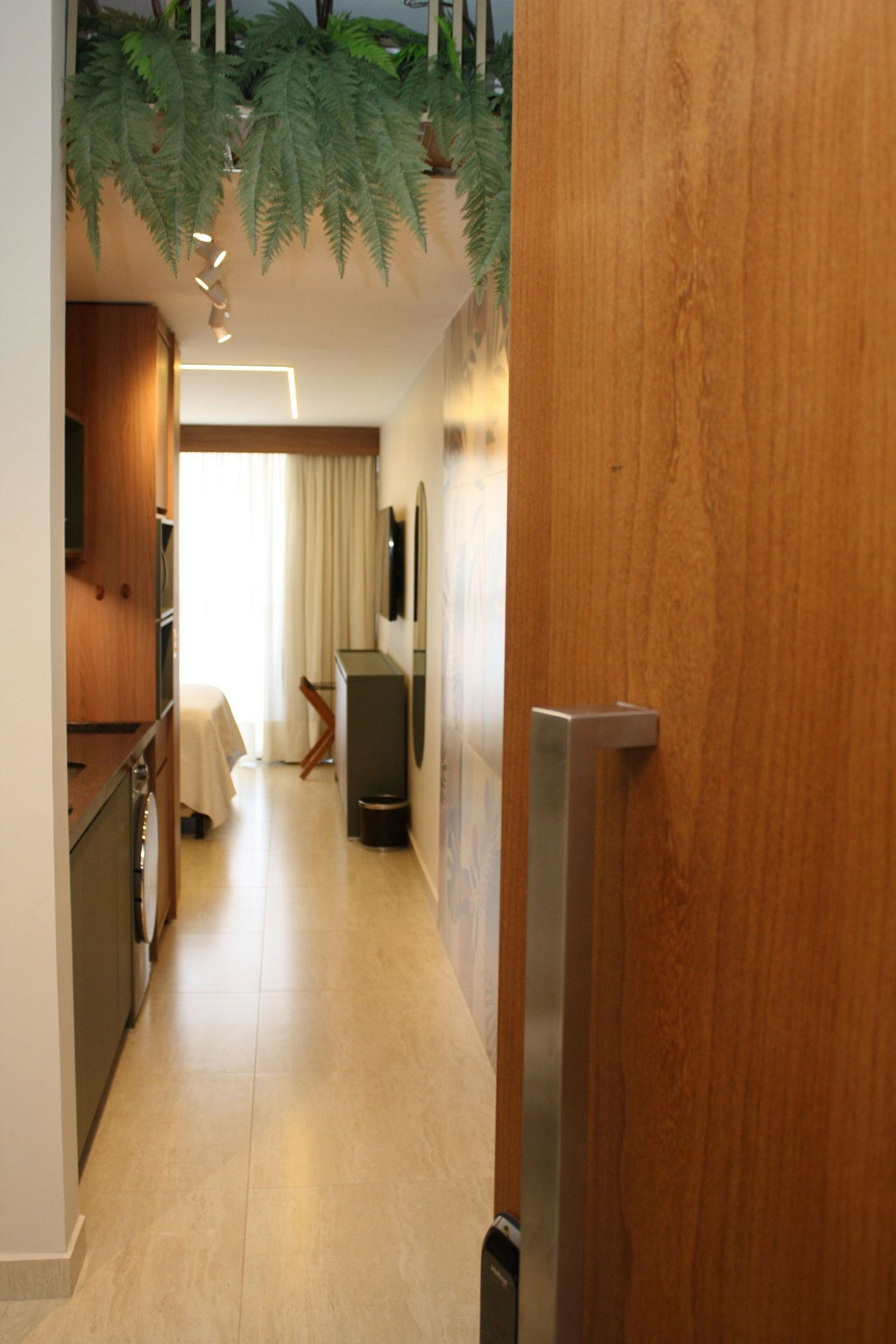 Un pasillo estrecho con una puerta de madera, una mesita con una silla, un televisor en la pared y una planta colgando del techo.