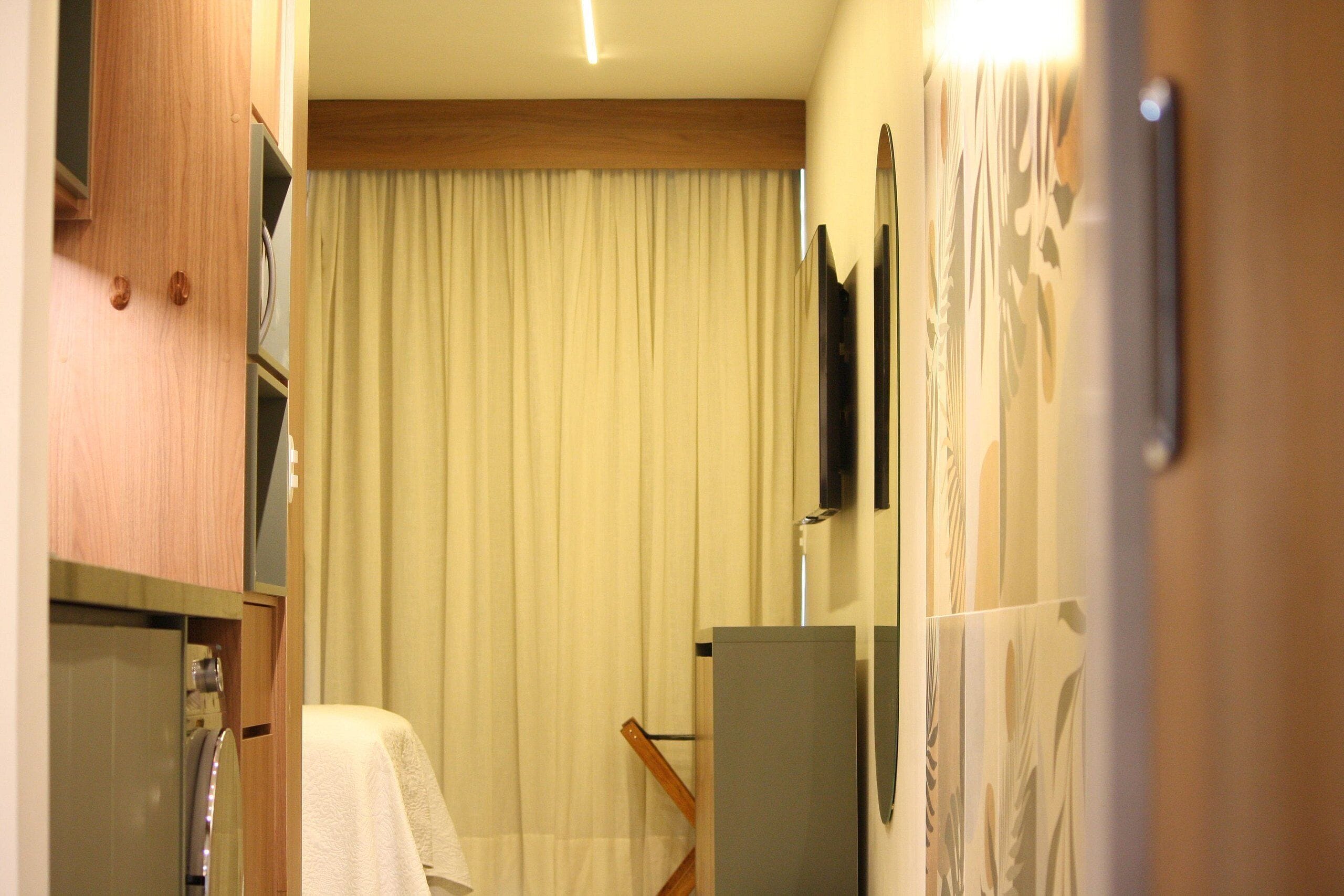 A narrow hotel room with a wooden wardrobe, a small table with a white cloth, a mirror, and a curtain.