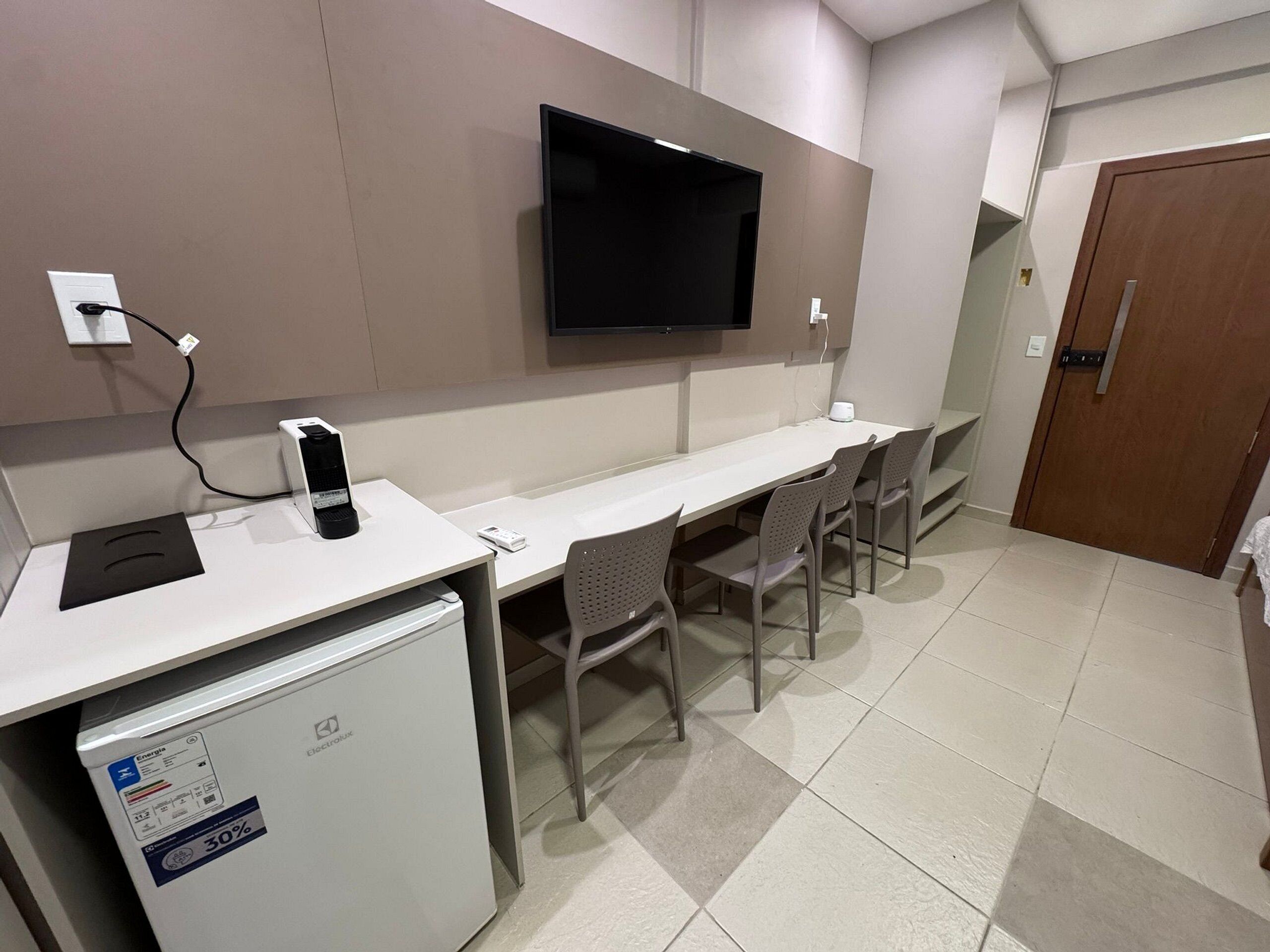 Ein kompaktes Hotelzimmer mit Küchenzeile, einem flachbildfernseher an der Wand, einem kleinen Kühlschrank und einer Essecke mit Stühlen.