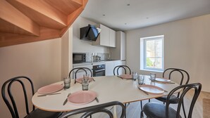 Un comedor con una mesa lista para cuatro personas, rodeada de sillas negras. Se ve una cocina con armarios blancos y un microondas al fondo.