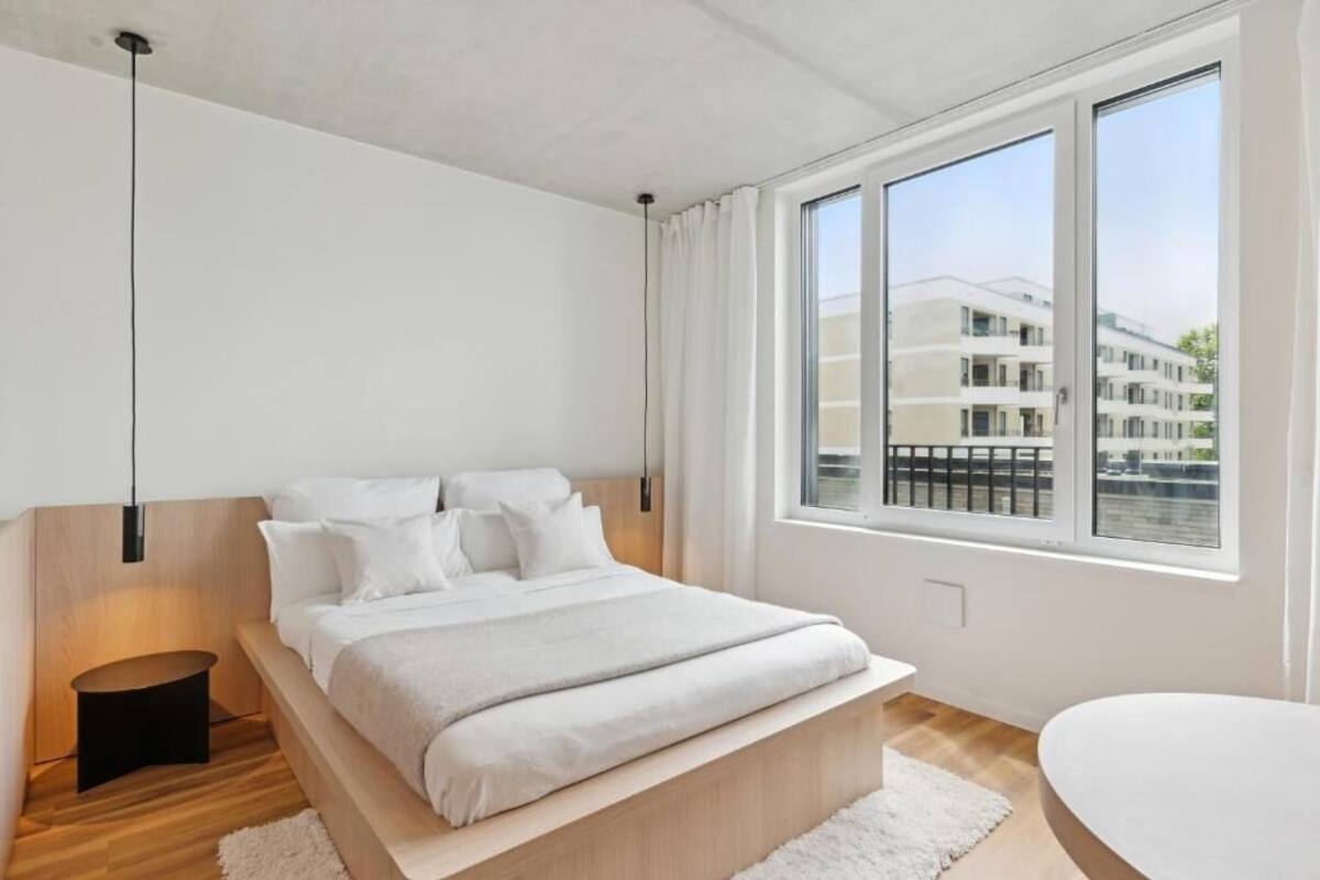 A modern bedroom with a large bed, a bedside table, a window with a view of buildings, and a white wall.