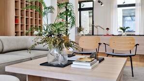 A modern office space with a wooden bookshelf, a sofa, two chairs, a table with a vase of plants, and books. - Habyt Berlin Waterfront (Berlin)