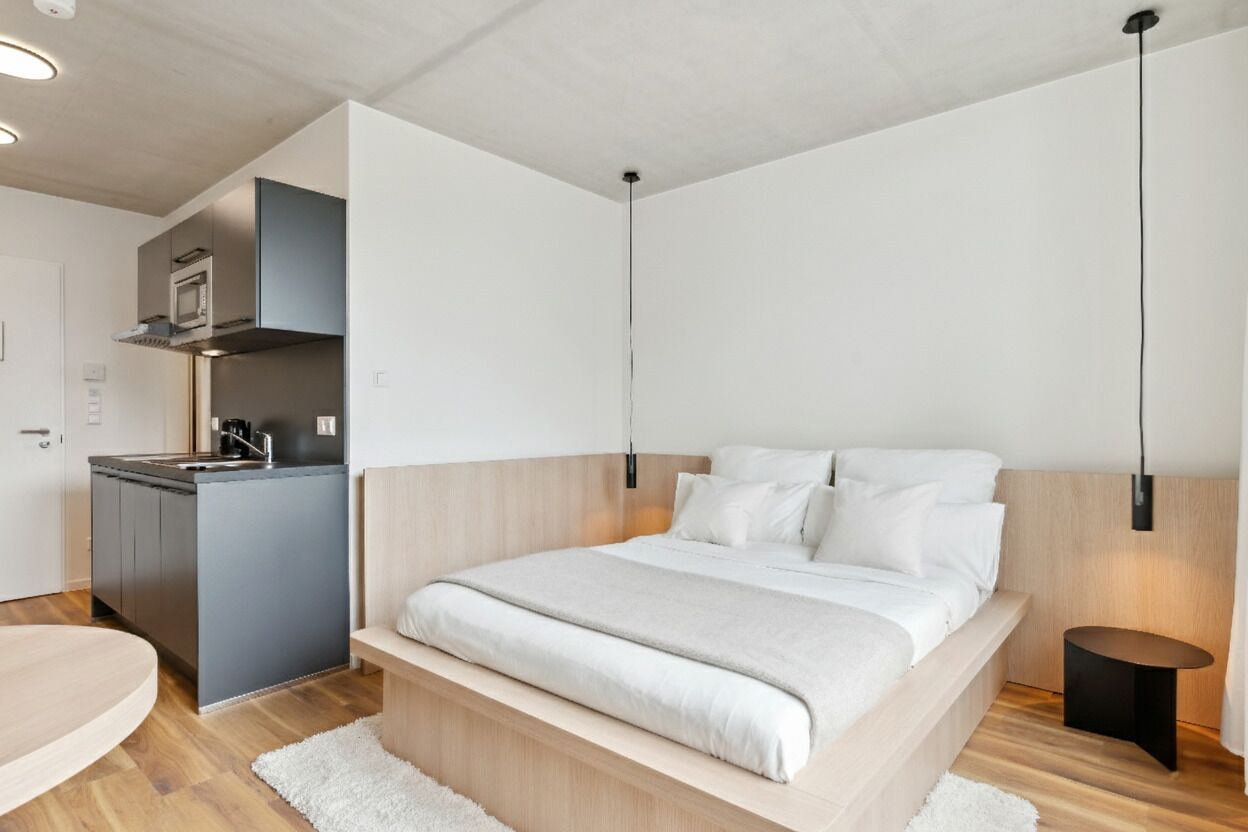 A modern bedroom with a wooden headboard, a small bedside table, and a built-in kitchen area with a microwave and stove.