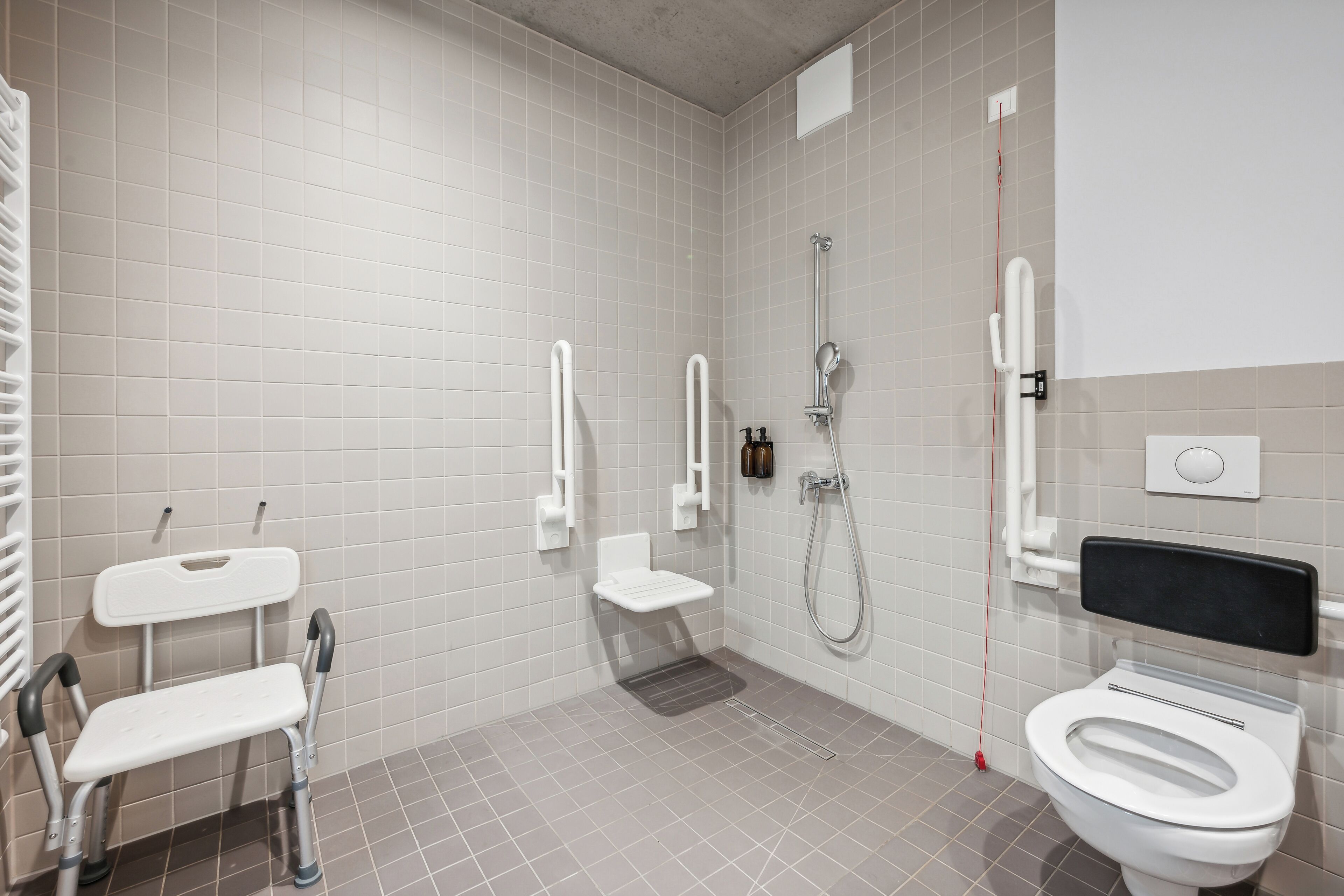 A modern bathroom with white tiles, a white toilet, a shower with a handheld showerhead, and a grab bar.