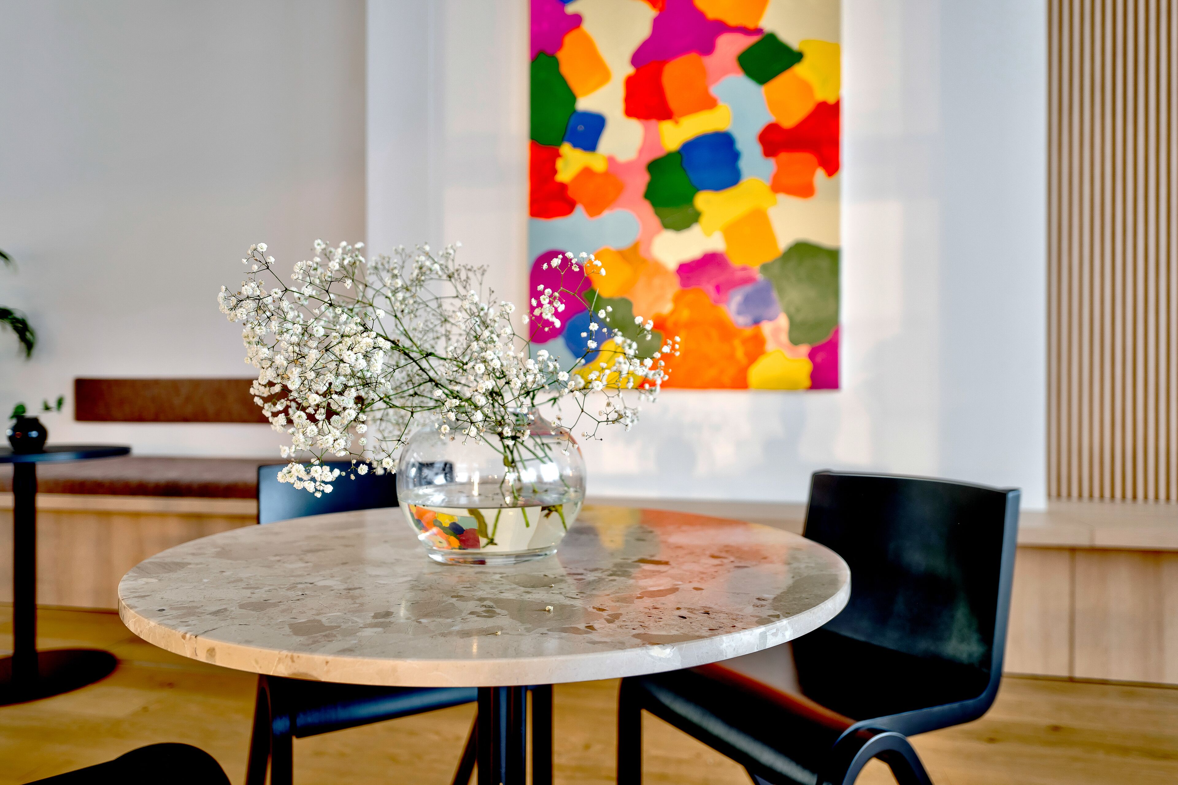 Una mesa redonda con un florero de flores blancas, un cuadro abstracto colorido en la pared y un banco de madera al fondo.