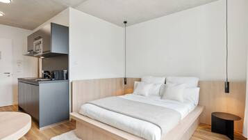 Un dormitorio moderno con cama de madera, una mesita de noche pequeña y una zona de cocina integrada.