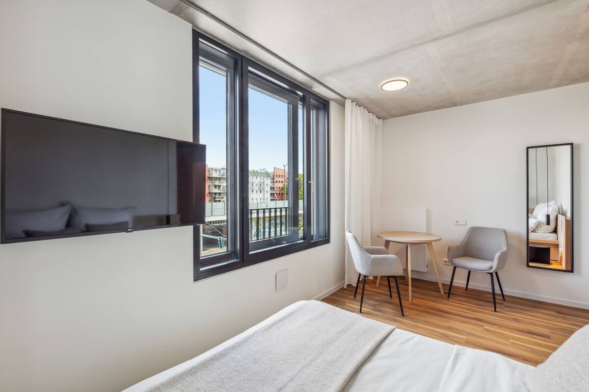 A modern hotel room with a large window, a bed, a desk, two chairs, and a mirror.