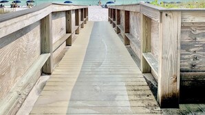Ein hölzerner Steg führt zu einem Strand mit blauem Himmel und ruhigem Wasser.