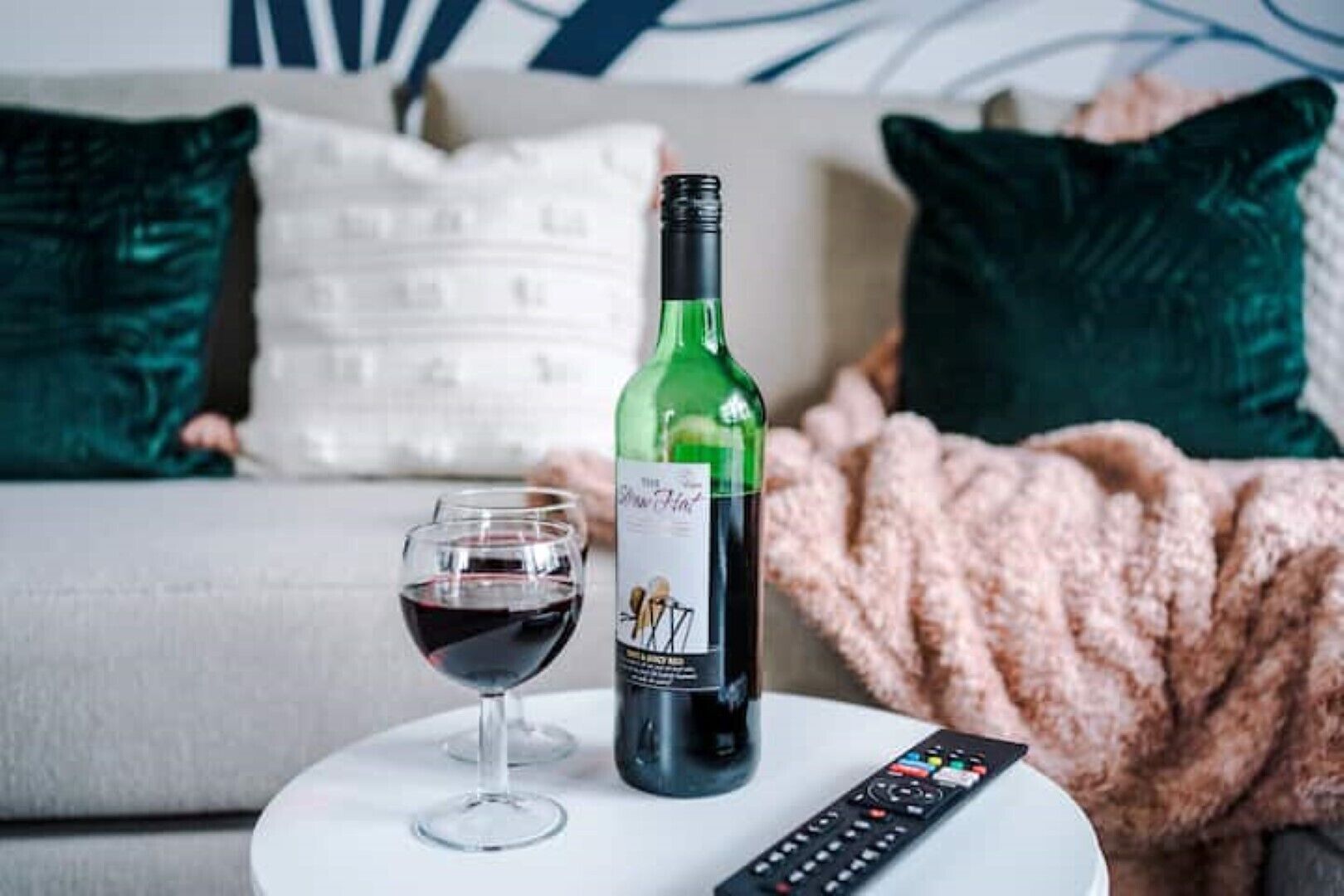 A bottle of wine and a glass on a white table in a living room with a couch and pillows in the background.