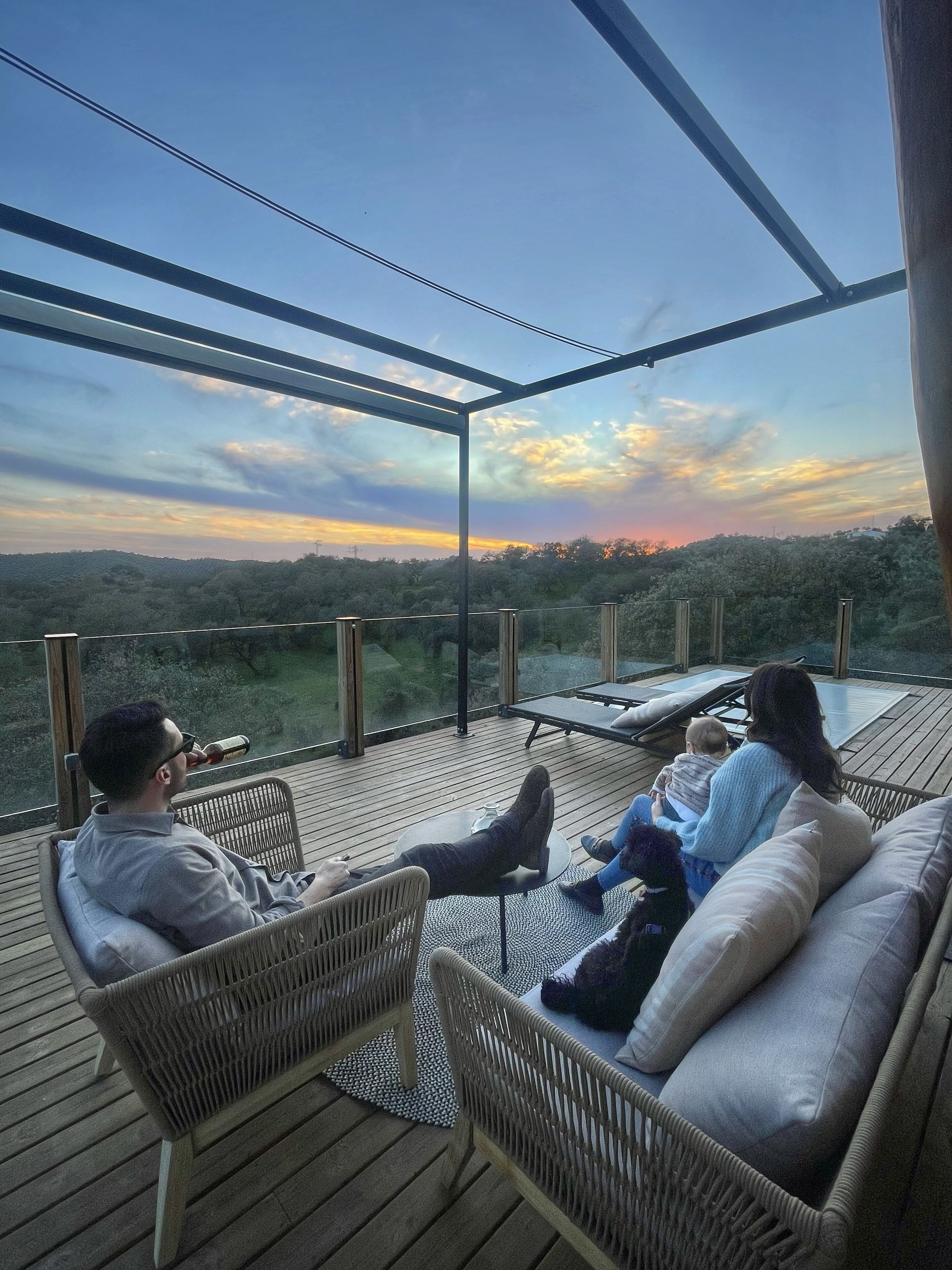 Una pareja y un niño descansando en una terraza de madera con techo de vidrio, con vista a un paisaje boscoso.
