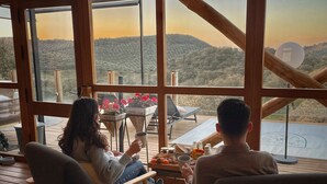 A couple seated at a table on a wooden deck with a view of a hilly landscape through large windows. - La Dehesa Experiences (Adamuz)