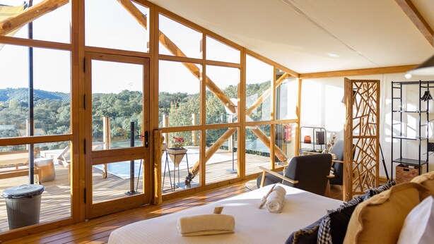 A spacious bedroom with a large bed, wooden floor, and a view of a deck and mountains. - La Dehesa Experiences (Adamuz)