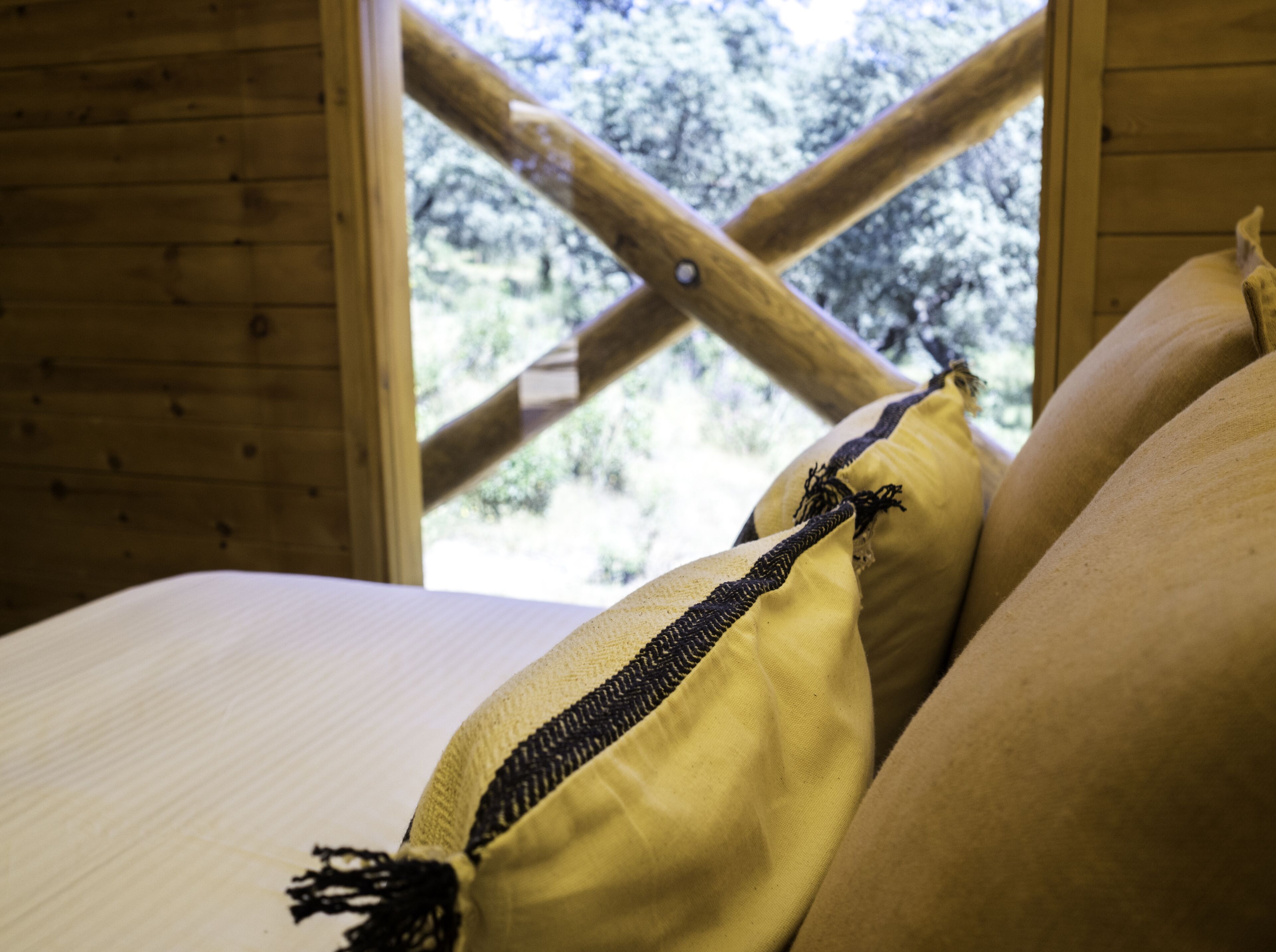 Interior de una cabaña de madera con una cama, almohadas y una ventana que da a un paisaje nevado.