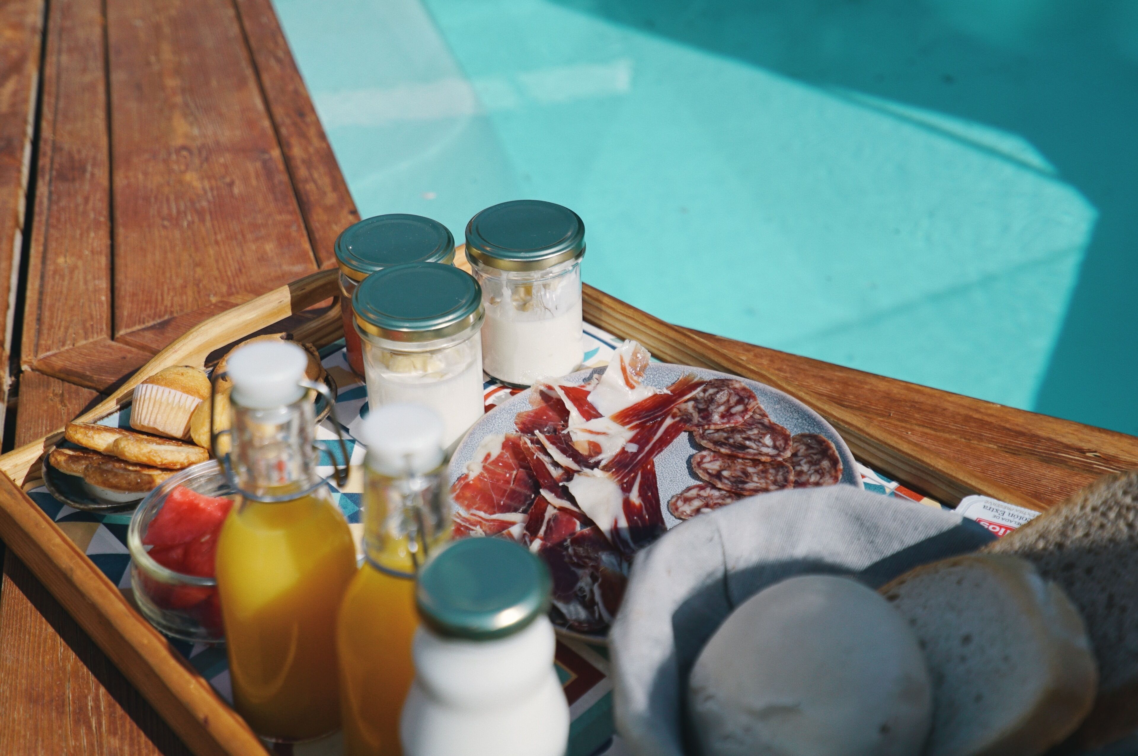 Una bandeja de madera con alimentos, incluyendo pan, un botellón de zumo de naranja y un plato de embutidos junto a una piscina.