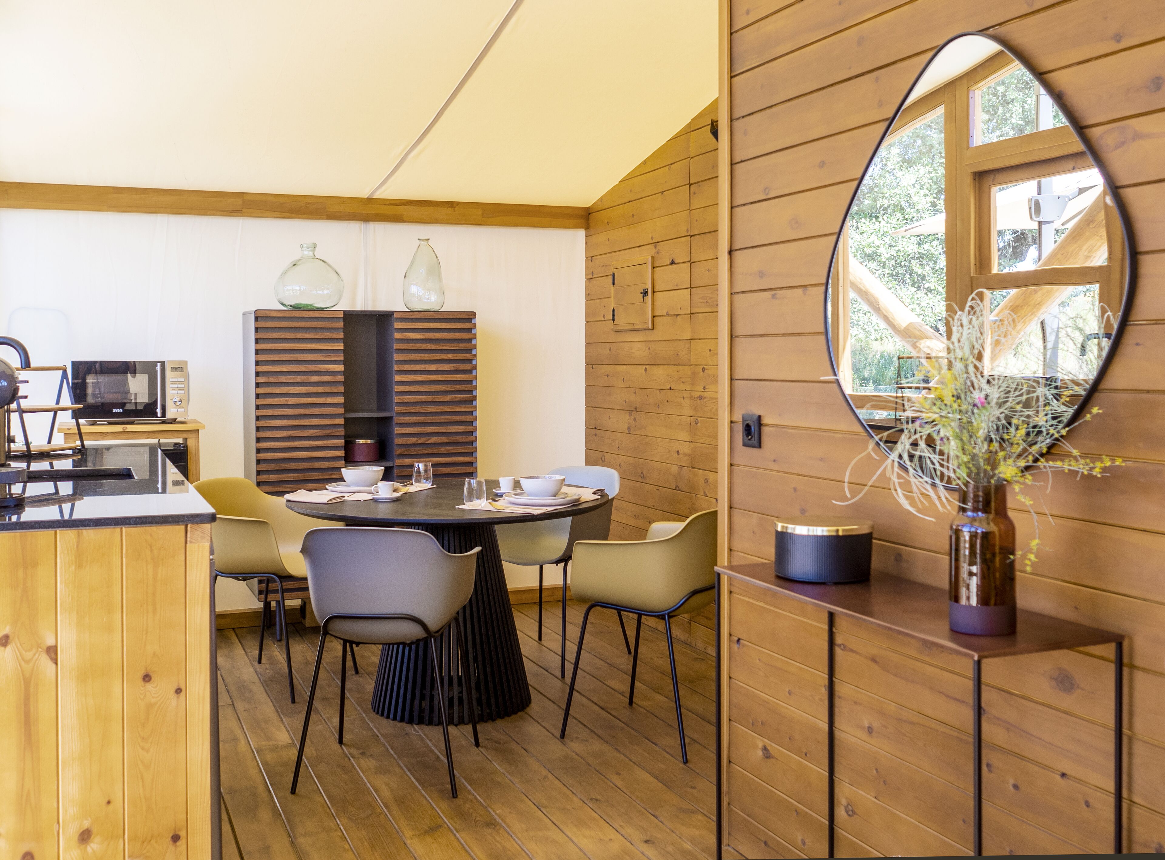 A modern dining area with a round table, chairs, a microwave, and a wooden cabinet with glass bottles.