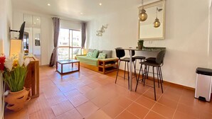 A modern living room with a sofa, coffee table, and dining area. There is a TV mounted on the wall, a window with curtains, and a potted plant. - ALBUFEIRA SKYLIGHT 1 WITH POOL by HOMING (Albufeira)