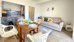 Un coin repas avec une table en bois, des chaises blanches et une cuisine en arrière-plan.
