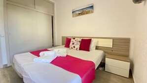 Un lit double bien fait, avec une couverture rouge et blanche, deux oreillers et une petite table de chevet surmontée d’une lampe.