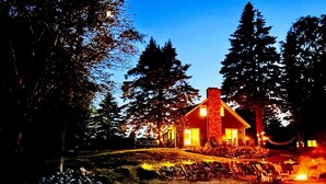 Un chalet confortable, doté d’une cheminée imposante, entouré d’arbres et comprenant un espace aménagé pour faire un feu de camp.