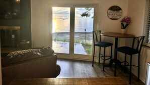 Un salon doté d’une grande porte-fenêtre coulissante donnant sur une terrasse avec vue sur l’eau. On y trouve un canapé, une petite table avec deux chaises et un panneau fixé au mur.