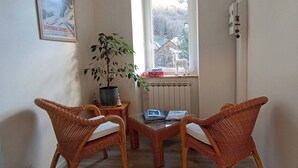 Een kleine kamer met twee rieten stoelen, een tafel, een potplant, een radiator en een raam met uitzicht op bomen en een hertenbeeld.