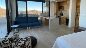 Una habitación de hotel moderna con balcón, un sofá, una mesita, un otoman decorado y una kitchenette.