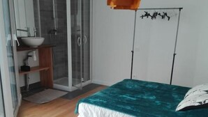 Une chambre avec un lit, une chaise, une petite salle de bain avec un lavabo et une douche, et une porte vitrée donnant sur un balcon.