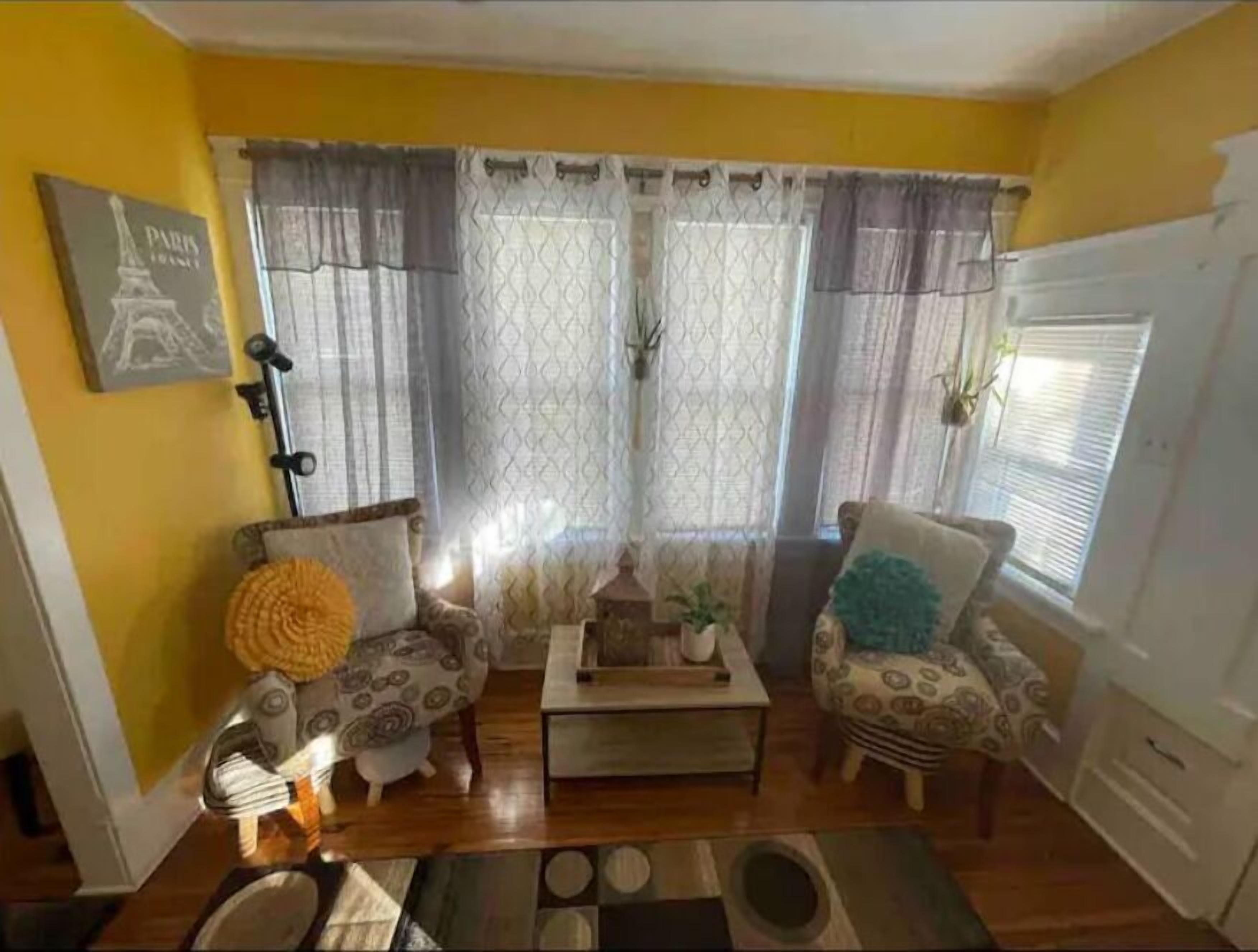 Een woonkamer met gele wanden, een houten vloer, twee gestreepte fauteuils, een salontafel en een decoratief kussen.