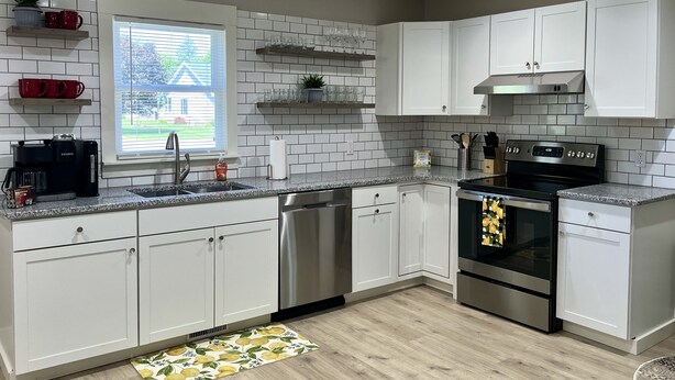 A modern kitchen with white cabinets, a stainless steel dishwasher, an oven, and a window with a view of a house and trees.