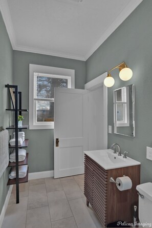 Ein Badezimmer mit einem weißen Waschbecken, einem Holzschrank und einem Spiegel. Eine Ablagefläche zeigt Handtücher und ein Fenster mit Jalousien.