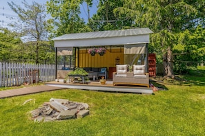 A small, modern outdoor structure with a white exterior and a metal roof, featuring a seating area with a sofa and a table, surrounded by greenery and trees. - Sailors Rest (Marblehead)