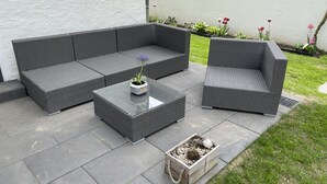 A patio area with a grey wicker sofa, a matching armchair, and a glass-top coffee table with a potted plant.