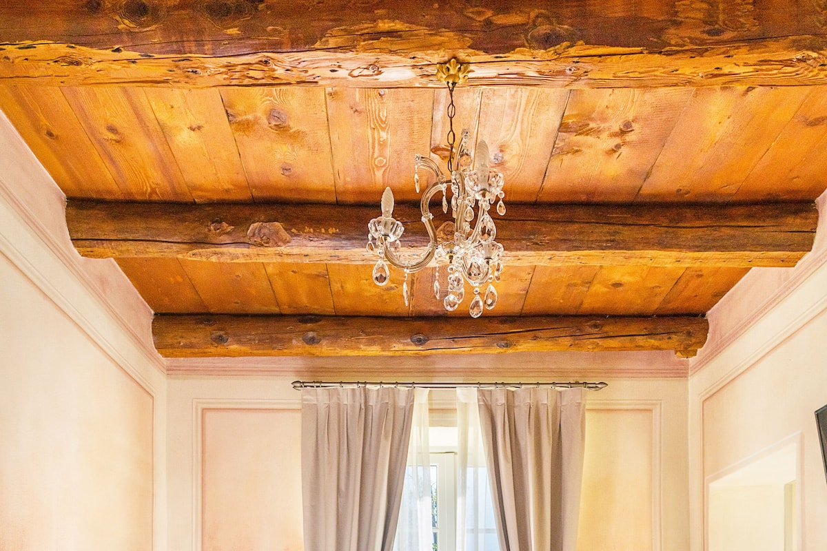 Une chambre avec un plafond en bois, un lustre, un lit avec une literie blanche et rose, une table de chevet et une fenêtre avec des rideaux.