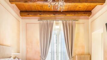 Un dormitorio con techo de madera, un candelabro, una cama con ropa de cama blanca y rosa, una mesita de noche y una ventana con cortinas.