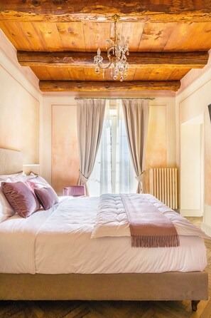A bedroom with a wooden ceiling, a chandelier, a bed with white and pink bedding, a bedside table, and a window with curtains. - VILLA CHIOVENDA Residenza d'Epoca (Premosello Chiovenda)