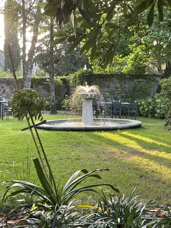 Un jardín con una fuente central, rodeado por un muro de piedra y abundante vegetación.