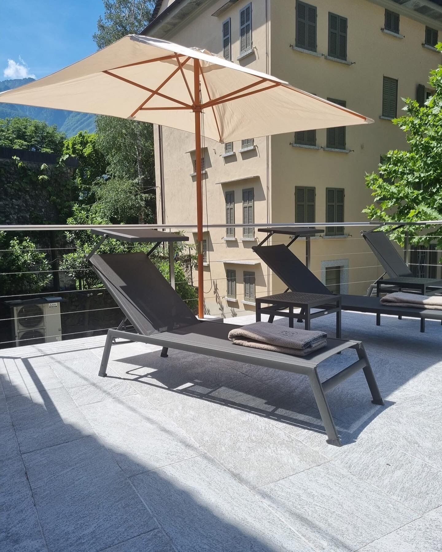 A rooftop terrace with two sun loungers, a beige umbrella, and a building in the background.