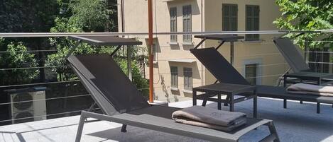 A rooftop terrace with two sun loungers, a beige umbrella, and a building in the background.