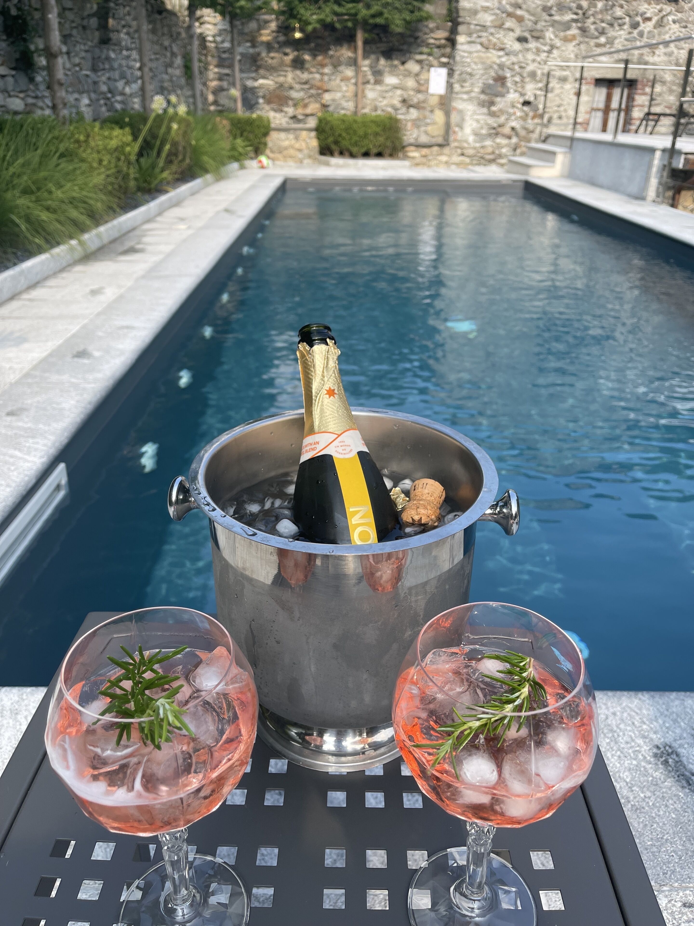 A poolside setting with a champagne bottle in a bucket, two glasses of pink cocktail, and a stone wall in the background.