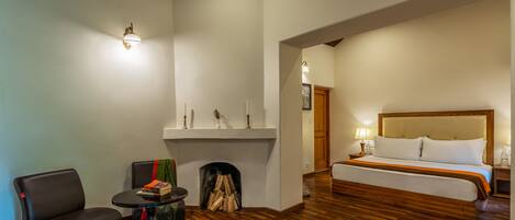 A hotel room with a wooden floor, a bed with white bedding, a small table with books, a chair, and a fireplace.