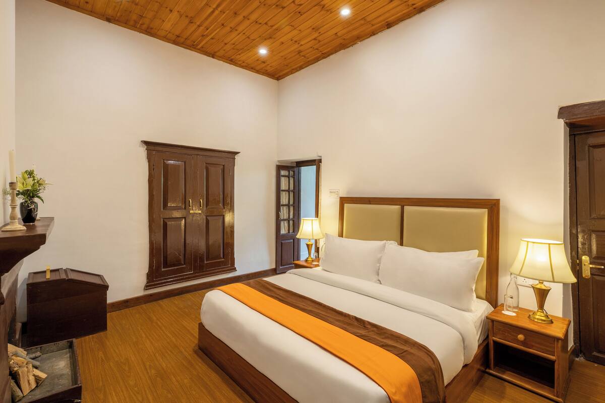 A hotel room with a wooden ceiling, a large bed with white and orange bedding, two bedside tables with lamps, a wooden wardrobe, and a small wooden chest.