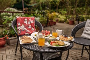 Una mesa servida con alimentos para el desayuno, como panqueques, croissants y una jarra de jugo de naranja, rodeada de sillas de mimbre y plantas en macetas.